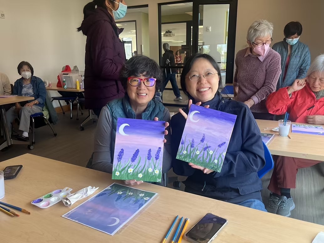 Two women smiling and holding up their lavender moon paintings at a CARES workshop