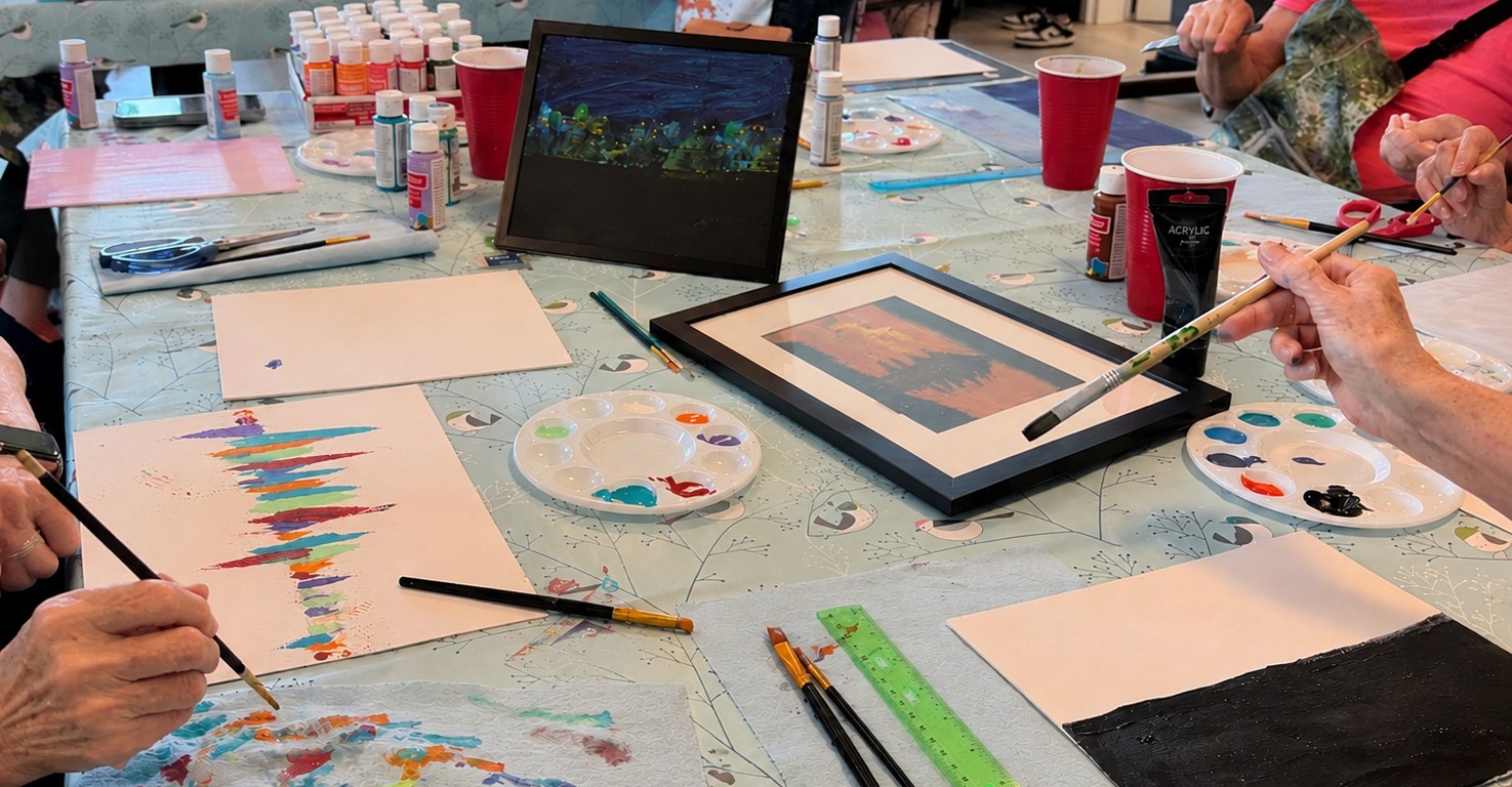 A workshop table covered in paintings in progress, paint palettes, brushes, and framed paintings, with residents painting