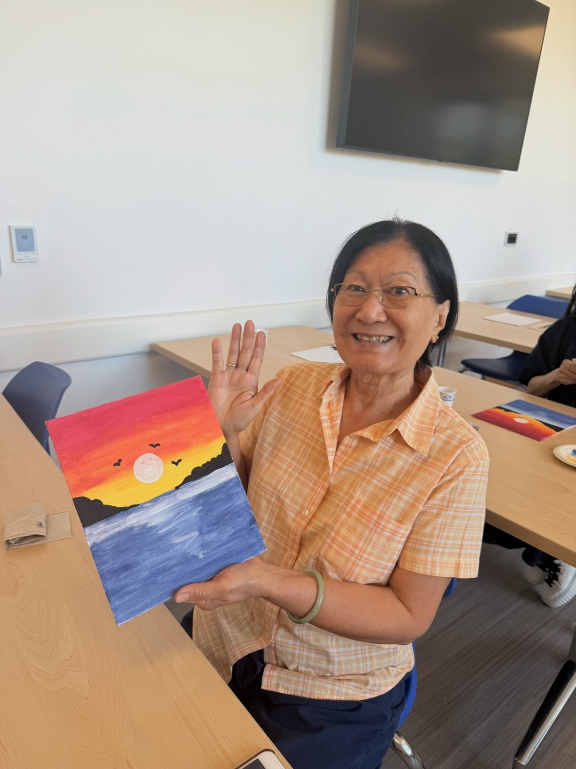 A participant in an orange top proudly holding up her sunset painting