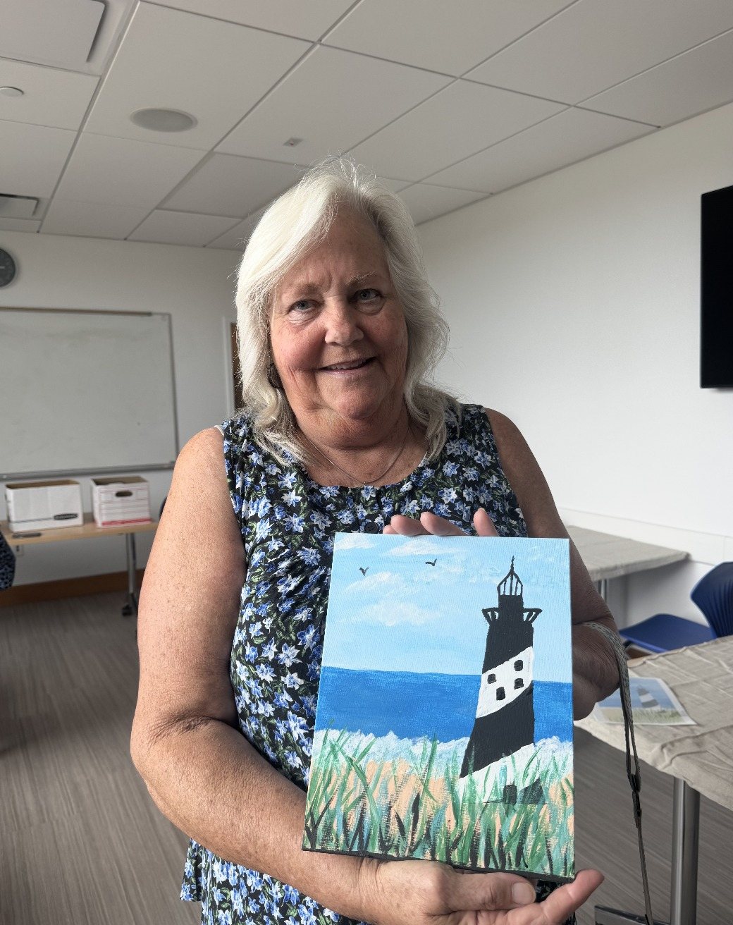A smiling participant holding up her painting of a striped lighthouse by the sea