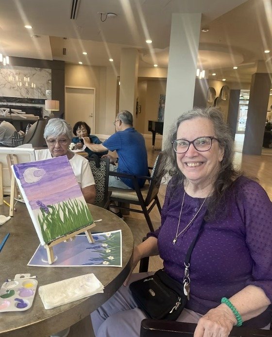 A smiling resident beside her painting of a moon over tall grass
