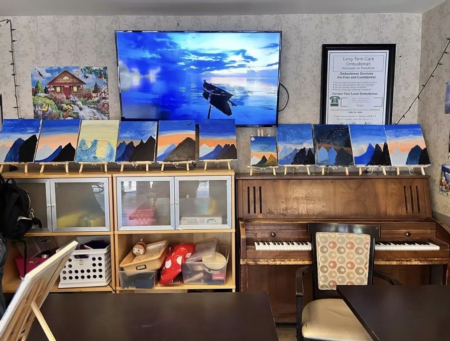 A row of finished mountain sunset paintings displayed on shelves near a piano